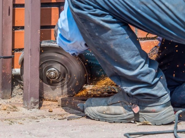 A gate technician wearing blue pants and black boots uses an angle grinder on a metal piece, creating sparks. The person is kneeling on the ground, with bricks visible in the background, likely preparing components for an electric gate installation.