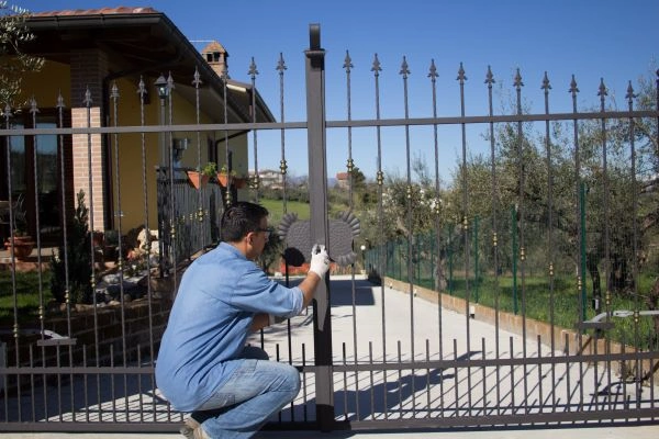 A man wearing a blue shirt and white gloves is crouching and working on a large, black wrought iron electric gate in front of a house in Houston. The gate is at the entrance to a paved driveway, with the house and surrounding landscape visible in the background under a clear blue sky.